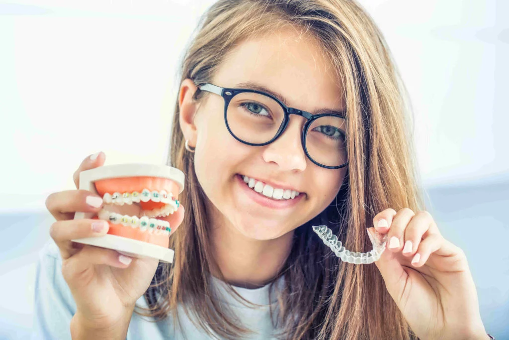illustration urgences Une adolescente portant des lunettes sourit tout en tenant un modèle dentaire avec un appareil dentaire dans une main et un aligneur orthodontique transparent dans lautre.