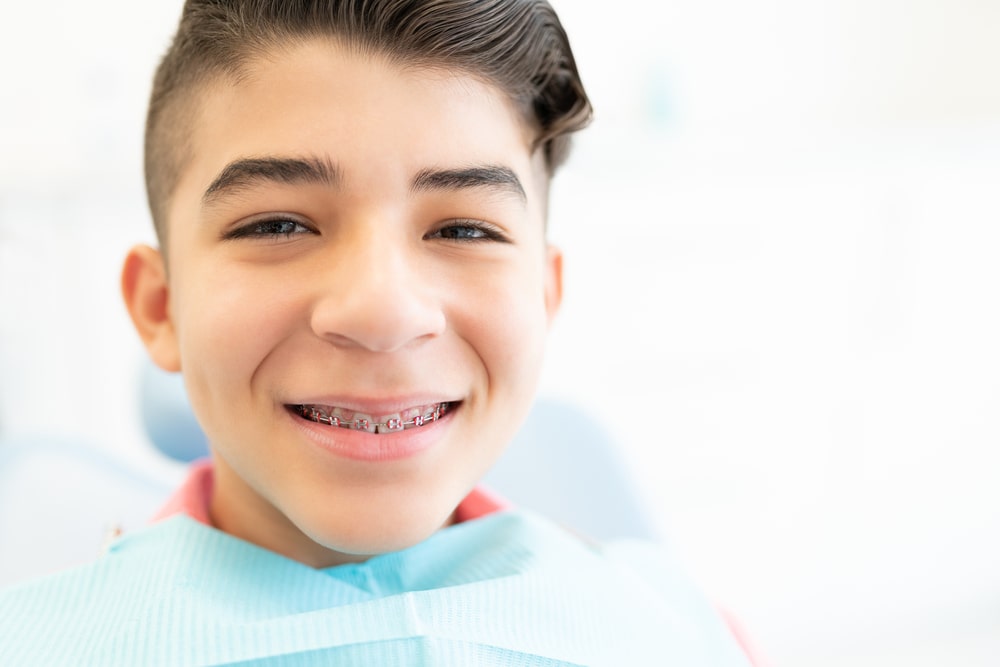 illustration arc blessant Un garçon souriant avec un appareil dentaire et un bavoir dentaire, assis sur une chaise de dentiste. Larrière-plan est lumineux et légèrement flou.