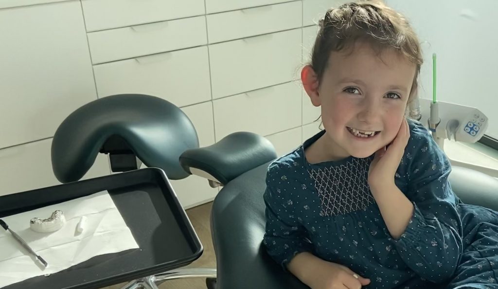 illustration urgences Un enfant assis sur un fauteuil de dentiste, portant une robe bleue à motifs floraux. À côté, une table roulante avec un modèle dentaire en plâtre et des outils dentaires. Le cabinet a des meubles blancs et un siège de dentiste vert foncé.